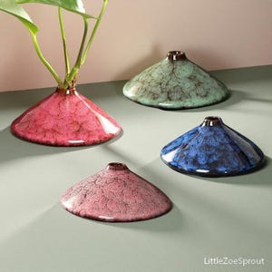 Puede incluir: Cuatro jarrones de cerámica en forma de cono en tonos rosa, verde y azul. Un jarrón contiene una planta con hojas verdes. Los jarrones tienen un esmalte texturizado y craquelado y un borde marrón oscuro. Los jarrones son decorativos.