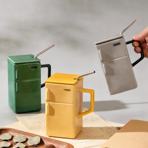 Op de afbeelding: Drie keramische mokken in de vorm van miniatuurkoelkasten, elk met een deksel, handvat en een metalen rietje. Eén is groen, één is geel en één is grijs. Op de gele mok staat het woord "FOCUS". De mokken staan op een licht oppervlak.