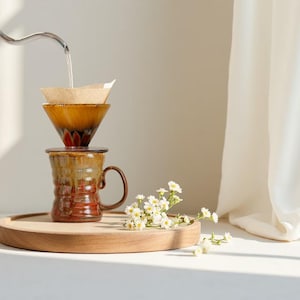Pode incluir: Um conjunto de café de cerâmica com esmalte marrom e vermelho, com um filtro de papel e uma caneca com alça. O conjunto está em uma bandeja de madeira, com flores brancas. A água está sendo despejada no filtro.