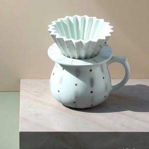 May include: A light blue ceramic pour-over coffee maker set. The set includes a mug with a handle and a matching filter holder. The filter holder has a unique, folded design. The mug and filter holder are decorated with small, dark gray dots.