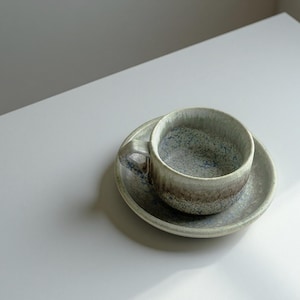 May include: A ceramic teacup and saucer set. The cup and saucer are a speckled gray with brown and blue accents. The cup has a handle and sits on a matching saucer, creating a minimalist aesthetic.
