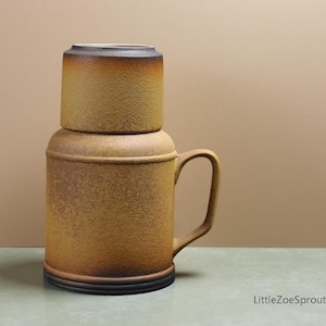 May include: A two-piece ceramic mug set in shades of brown and tan. The larger mug has a handle and a textured surface, with a smaller, cylindrical lid on top. The base is dark brown.