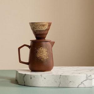 May include: A brown ceramic coffee pour-over set. The set includes a carafe with a handle and a lid, and a cone-shaped filter holder. The carafe has a speckled design. The filter holder has a light brown and white rim.