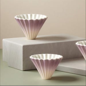 Puede incluir: Tres vertedores de café de cerámica con un diseño acanalado y una combinación de colores degradados de blanco a lavanda. Los vertedores en forma de cono están sobre una superficie gris claro, sugiriendo una estética minimalista.