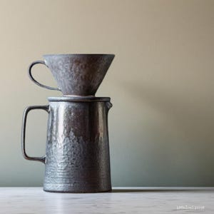Może przedstawiać: Ciemnoszary ceramiczny zestaw do parzenia kawy metodą pour-over. Zestaw zawiera teksturowany, stożkowy uchwyt na filtr i pasujący dzbanek z uchwytem. Zestaw do kawy jest przeznaczony do parzenia kawy i ma rustykalny, ręcznie robiony wygląd.