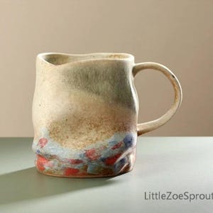 May include: Handmade ceramic mug with a unique, textured design. The mug features a cream-coloured body with a green and brown gradient, and a colourful base with red and blue accents. The handle is curved and matches the body's colour. A functional and decorative piece.