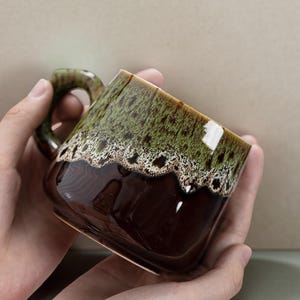 Może przedstawiać: Ceramiczny kubek o kwadratowym kształcie i zaokrąglonym uchwycie. Kubek ma ciemnobrązową podstawę, przechodzącą w zielono-brązowy, nakrapiany wierzch. Obręcz ma biały, teksturowany wzór. Kubek trzymany jest w dwóch dłoniach.