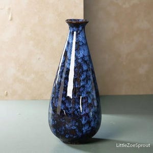 May include: A tall, blue ceramic vase with a glossy finish. The vase has a narrow neck and a wider, rounded body. The color transitions from dark blue at the base to a lighter blue with a speckled pattern towards the top. The rim is a dark brown.
