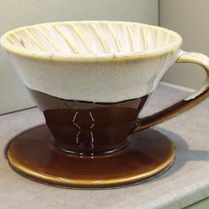 May include: A ceramic pour-over coffee maker with a brown, cream, and tan color scheme. The coffee maker includes a cup, a handle, and a saucer. The top portion is cream-colored, the middle is brown, and the bottom saucer is brown.