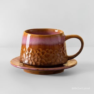 May include: A ceramic mug and saucer set. The mug has a textured, bubble-like pattern on the lower half and a smooth, glossy finish on the upper half. The colors transition from a deep brown to a pinkish-purple. The saucer is a matching color.