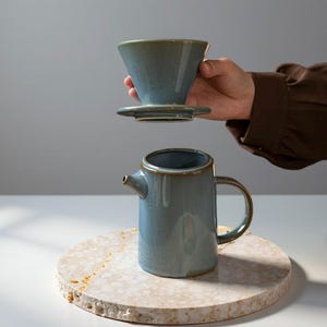 May include: A blue ceramic pour-over coffee maker set. The set includes a cone-shaped dripper, a base with a handle, and a small spout. The set is on a round, light-coloured stone surface.