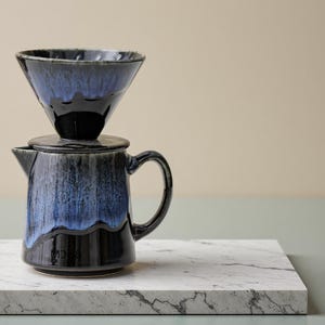 Pode incluir: Um conjunto de cafeteira de cerâmica para servir. O conjunto inclui um gotejador em forma de cone e uma jarra a condizer. O gotejador e a jarra são esmaltados num esquema de cores azul escuro e preto, com um efeito de gotejamento. O conjunto está numa superfície de mármore.