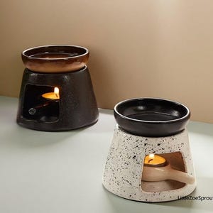 May include: Two ceramic oil warmers. One is black with a brown top, the other is white with black speckles and a black top. Each has a lit candle inside, visible through a rectangular opening.