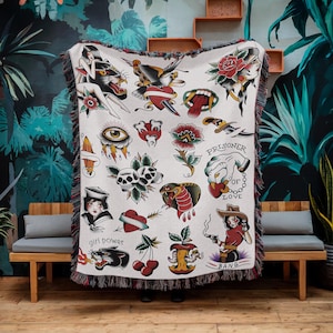 Puede incluir: Manta crema con diseños de tatuajes tradicionales. La manta muestra rosas, serpientes, calaveras y una vaquera con el texto "BANG". La manta tiene un borde con flecos. Los diseños son en rojo, negro y verde.