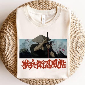 May include: A cream-colored crew neck t-shirt featuring a rectangular graphic of a warrior in a conical hat holding a sword, with red Japanese kanji text printed below the image. The shirt has a standard short-sleeve design and a round neckline.