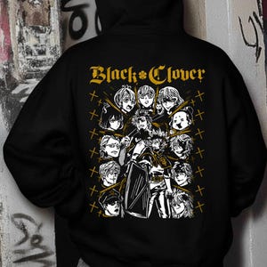 May include: A black hooded sweatshirt featuring a large graphic on the back. The design includes the text Black Clover in gold, surrounded by a collection of black and white anime character illustrations and small gold cross symbols arranged in a grid.