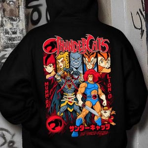 Könnte beinhalten: Ein schwarzer Kapuzenpullover mit einem großen Grafikdruck auf der Rückseite. Das Design zeigt das ThunderCats-Logo, Charaktere wie Lion-O und Mumm-Ra sowie Text auf Englisch und Japanisch. Die Illustration verwendet leuchtende Rot-, Blau- und Gelbtöne.