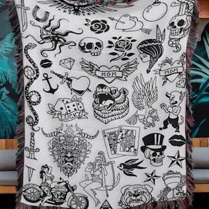 May include: A white woven throw blanket featuring a black ink-style print of various traditional tattoo designs, including skulls, roses, a bear, a motorcycle, playing cards, an anchor, and banners with the text MOM and LUCKY.