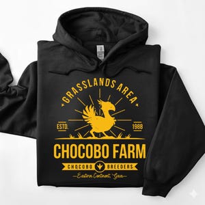 May include: Black hooded sweatshirt with a yellow graphic. The graphic includes the text "Grasslands Area," "Chocobo Farm," and "Chocobo Breeders." A stylized yellow bird is in the center of the design, with the year "1988" and the text "Eastern Continent, Gaia."