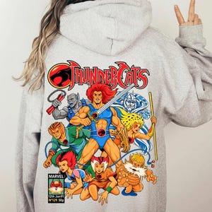 May include: A light grey hooded sweatshirt featuring a large, colourful graphic on the back depicting the Thundercats characters in action. The design includes the red Thundercats logo, a Marvel comic book box labelled #129 Jan91, and various figures.