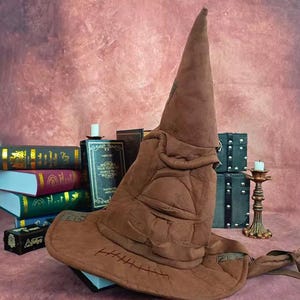 May include: A brown, pointed wizard's hat with a wide brim and stitched details. The hat is resting on a stack of books, with a candle and decorative boxes in the background. The hat's design is reminiscent of a classic sorting hat.