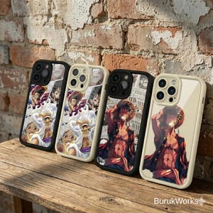 May include: Four phone cases with vibrant anime character designs. The cases have black and cream-coloured frames. The designs feature various characters in action poses. The cases are displayed on a wooden surface.