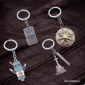 Op de afbeelding: Vier metalen sleutelhangers met zilveren sleutelringen. Eén sleutelhanger heeft een rechthoekig label met de tekst "PRAISE THE SUN". Een andere is een cirkelvormig medaillon met een zonontwerp en de tekst "PRAISE THE SUN". De andere twee hebben game-gerelateerde ontwerpen.