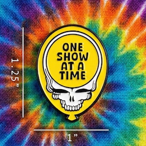 May include: A yellow enamel pin in the shape of a balloon, featuring a white skull design and the text "ONE SHOW AT A TIME" in black. The pin measures 1.25 inches high and 1 inch wide. The background is a colorful tie-dye pattern.