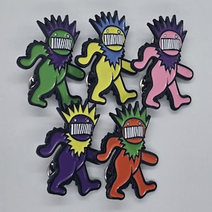 May include: Five colourful enamel pins depicting a stylized bear design. The pins are in green, yellow, pink, purple, and orange, each with a black outline, jagged collar, and a white-toothed grin.
