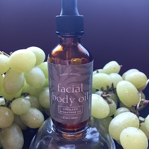 Puede incluir: Una botella de vidrio marrón de aceite facial y corporal con un cuentagotas negro, etiquetada como "grapes skincare". La etiqueta dice "facial body oil" con "oregano grapeseed oil". La botella está rodeada de uvas verdes.