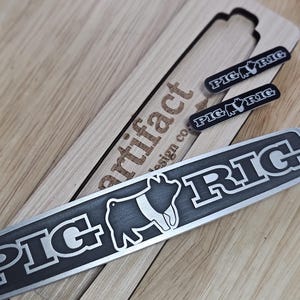 May include: A wooden box with a cutout for a metal plaque that reads "Pig Rig". The plaque features a stylized pig design. The box also has the text "Artifact Design Co." engraved on the side.
