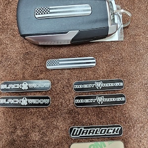 May include: A black car key fob with an American flag design.  The key fob is surrounded by several small black and white stickers with various text, including "Black Widow", "Rocky Ridge", and "Warlock".