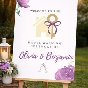 May include: A white sign on a wooden easel, announcing a housewarming ceremony. The sign has text reading "Welcome to the House Warming Ceremony of Olivia & Benjamin." It's decorated with purple flowers and a gold key graphic.