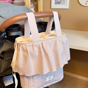 Könnte beinhalten: Eine rosa-weiß karierte Kinderwagen-Organizer-Tasche mit Rüschenbesatz. Die Tasche hat zwei Griffe und ist an einem Kinderwagen befestigt. Der Text "TinyZip Handmade" ist im Hintergrund sichtbar.