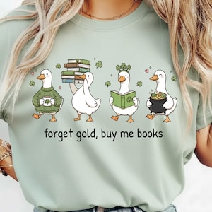 May include: Sage green t-shirt with a cartoon illustration of four ducks, each with a St. Patrick's Day theme. The ducks are shown with books, a pot of gold, and shamrocks. The text "forget gold, buy me books" is printed below the ducks.