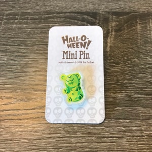 May include: Green gummy bear with a skull and crossbones design on its chest. The gummy bear is on a white card with the text "Hall-O-Ween! Mini Pin" and "Hall-O-Ween! © 2018 Tia Perkin".