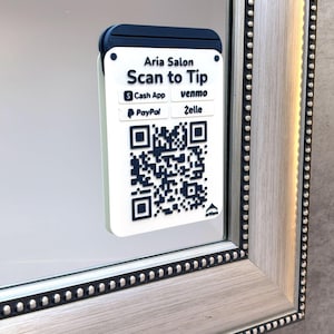 May include: A white and navy blue QR code sign that says "Aria Salon Scan to Tip" with payment options. The sign is attached to a silver framed mirror.