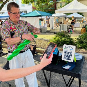 May include: A man in a colorful shirt twists a green balloon. A person holds a phone up to a sign that says "Pete K Scan to Tip" with QR codes for payment apps. The scene takes place at an outdoor market.