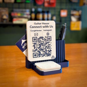 May include: A navy blue desk organizer with a white vertical sign featuring the text Guitar House Connect with Us, icons for Google Review, Instagram, email, and a website, plus a large QR code. It holds pens and a stack of white cards.