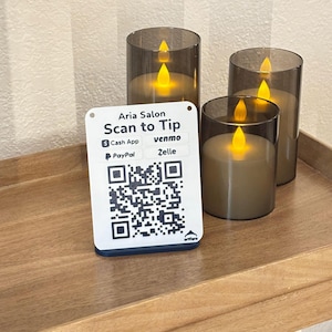 Puede incluir: Tres velas que funcionan con pilas con un efecto de llama parpadeante se exhiben en una bandeja de madera. Un letrero blanco con el texto "Aria Salon Scan to Tip" y un código QR está frente a las velas. El letrero también enumera opciones de pago.