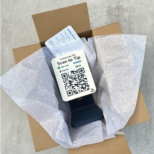 May include: An open cardboard box containing a Guitarman Kit. Inside, a white card with a QR code and the text "Scan to Tip" is displayed, along with a dark blue stand. The box also includes a "Getting Started" card and is lined with white tissue paper.