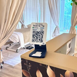 May include: Aria Salon Scan to Tip sign with QR codes for Cash App, Venmo, PayPal, and Zelle. The sign is white with a navy blue base, placed on a gold and black counter with floral designs. A massage table and curtains are in the background.
