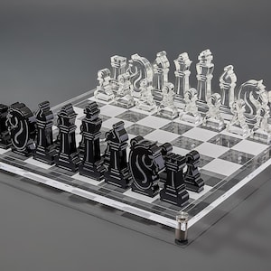 May include: A clear acrylic chess set with black and clear pieces. The chess board has a checkered pattern. The pieces are arranged on the board, ready for play. The set is displayed on a gray surface.