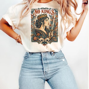 May include: Cream-colored t-shirt featuring a detailed graphic. The graphic depicts a woman wearing a crown and holding a sword, with the words "NO KINGS" above her. The design includes floral elements and two skulls at the bottom.