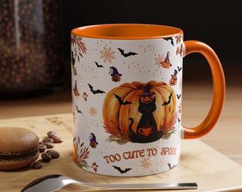 May include: White ceramic Halloween mug with an orange interior and handle. The mug features a black cat sitting on a pumpkin, surrounded by bats, spiders, and the text "TOO CUTE TO SPOOK".