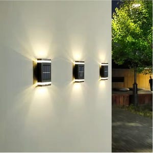 May include: Three black solar-powered wall lights with a rectangular design are mounted on a light-colored wall. Each light has a solar panel and emits a warm, downward-facing glow. The lights are evenly spaced.
