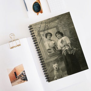 May include: A vintage black and white photo of two women standing in front of a window. The women are wearing dresses and holding flowers. A white dog is sitting in front of them. The photo is framed and has a handwritten message on the back that says "amigas que la vida te envia" and "Julia Guillermo".