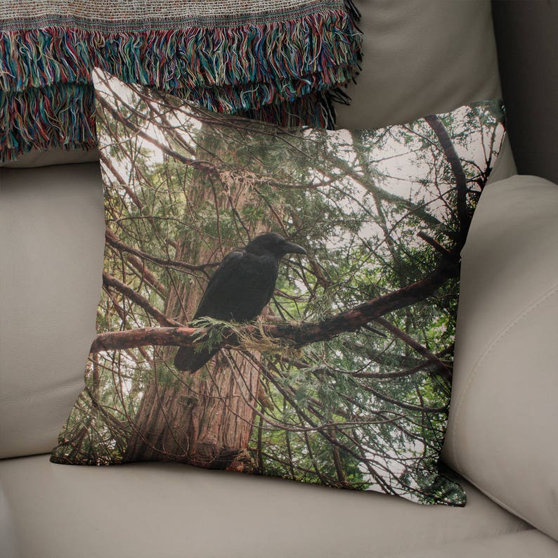 May include: A black crow perched on a branch in a forest. The crow is facing left and has its wings folded. The background is a blurred image of green trees.