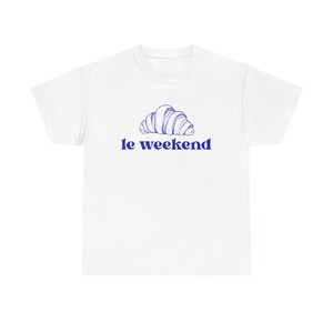 Puede incluir: Camiseta blanca con una ilustración azul de un cruasán y el texto "le weekend" en azul. El diseño está centrado en la parte delantera. Esta camiseta es una prenda casual para el uso diario.
