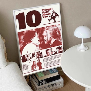 May include: A framed poster with a red and white design featuring the movie title "Things I Hate About You" and images of the cast. The poster is propped up against a wall, with books and a lamp visible.